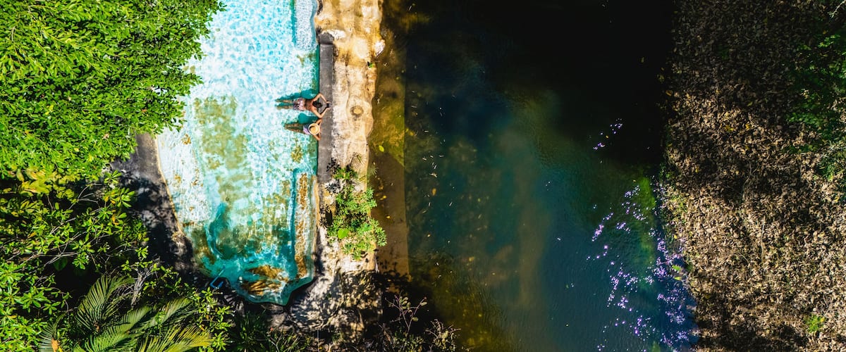 Surrounded by lush greenery, two individuals enjoy a peaceful moment by a pool that flows into a calm river. The vibrant colors of nature create a perfect tropical retreat for relaxation in Thailand