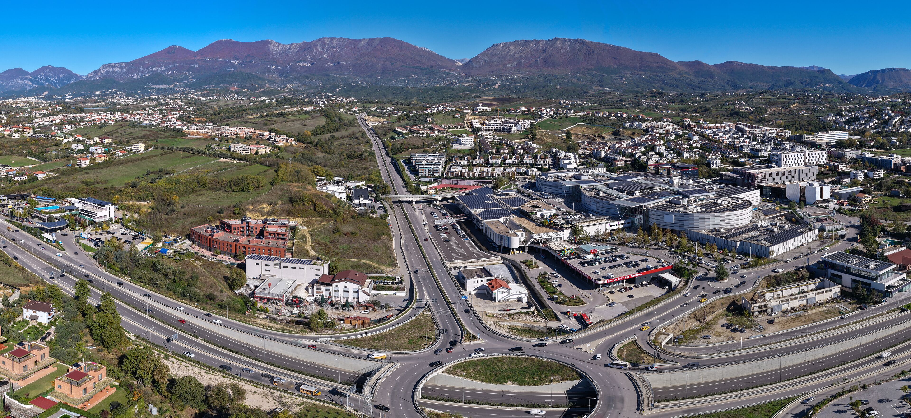 Panoramic aerial view of Tirana East Gate with highway, roundabout, shopping centre, and Sauk–Farkë suburbs surrounded by mountain landscape.