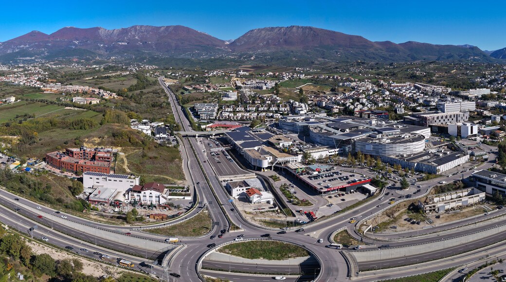 Panoramic aerial view of Tirana East Gate with highway, roundabout, shopping centre, and Sauk–Farkë suburbs surrounded by mountain landscape.