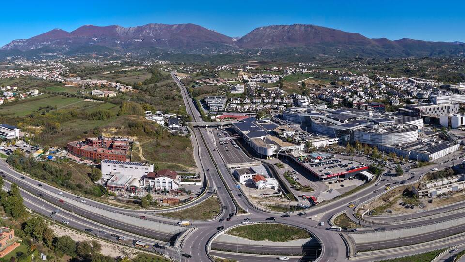 Panoramic aerial view of Tirana East Gate with highway, roundabout, shopping centre, and Sauk–Farkë suburbs surrounded by mountain landscape.