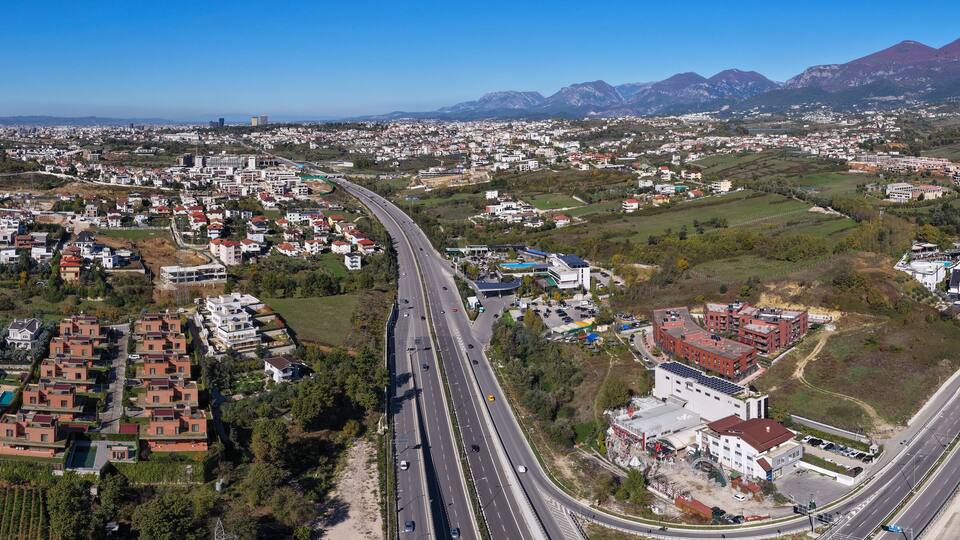 Panoramic aerial of Tirana’s eastern suburbs showing the highway, Sauk–Farkë residential area, and mountain landscape under clear skies.