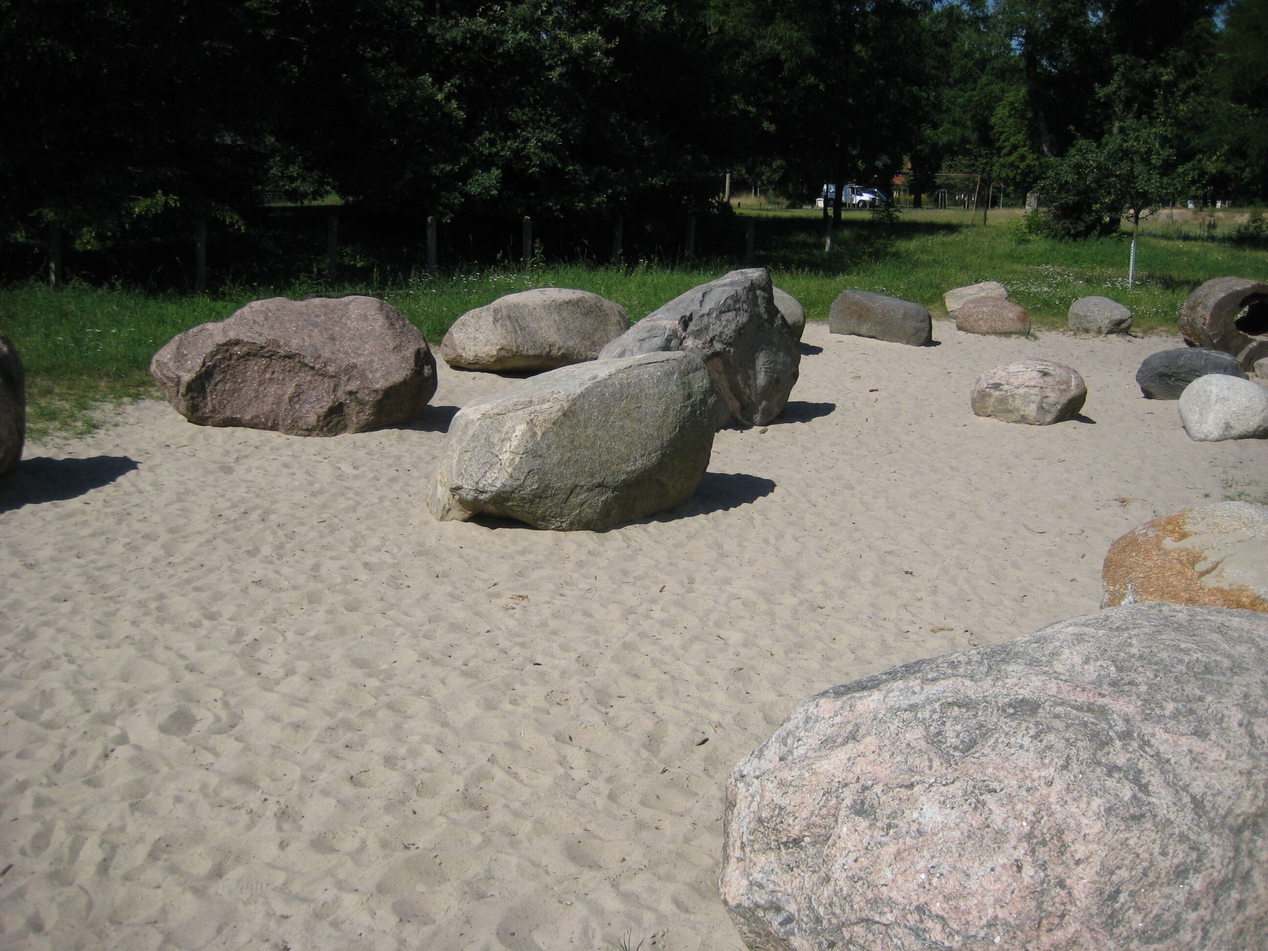 A sandy area with large rocks and a grassy field with trees in the background.