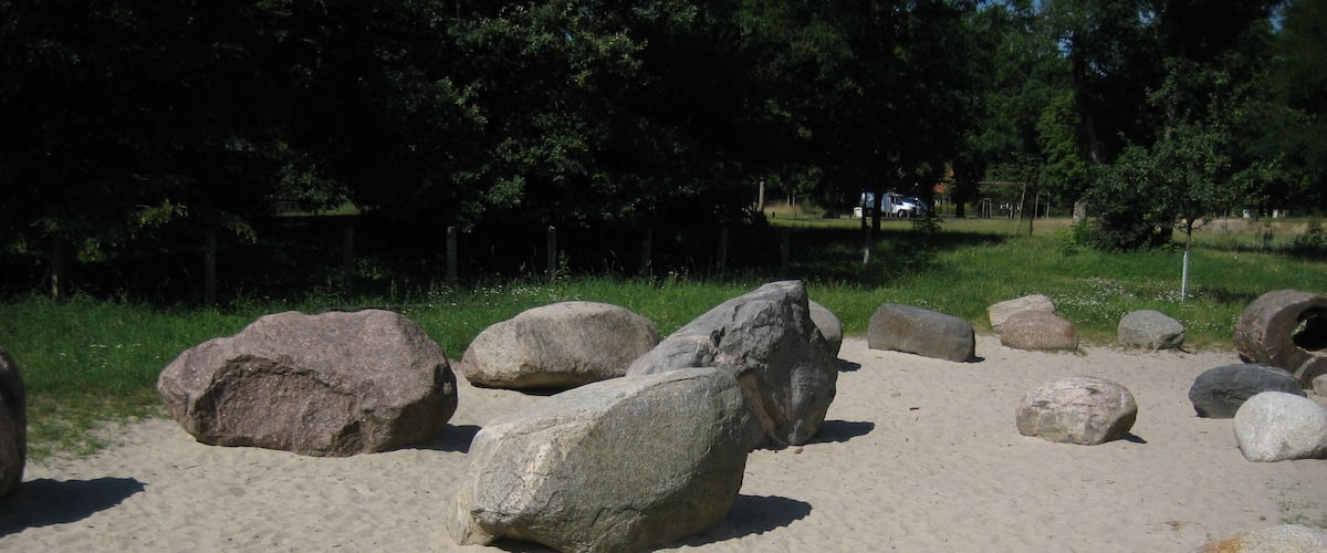 A sandy area with large rocks and a grassy field with trees in the background.