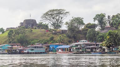 Uma vila à beira-rio com casas coloridas, uma grande fortaleza numa colina e vegetação exuberante.