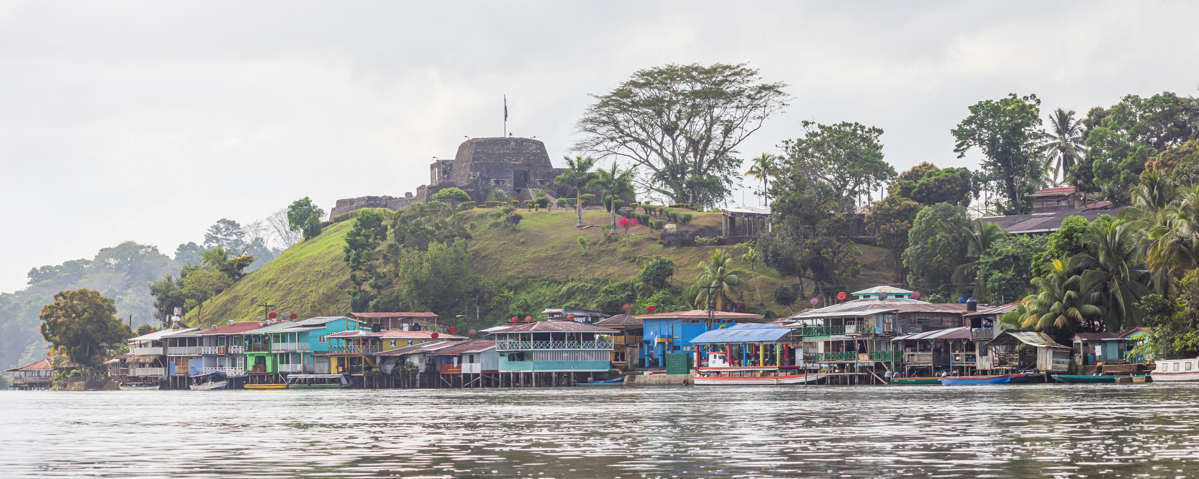 A riverside village with colorful houses, a large fort on a hill, and lush greenery.