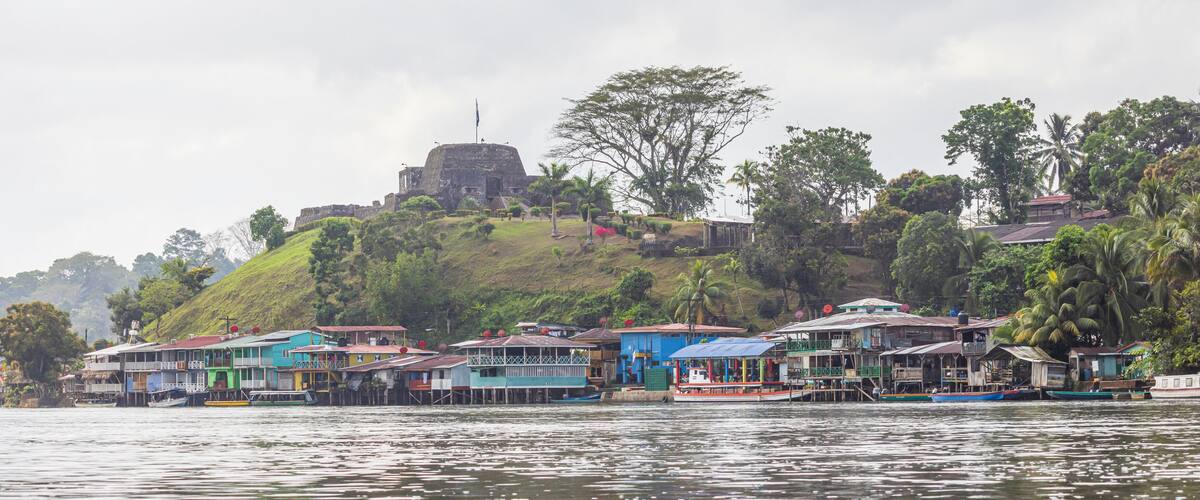 A riverside village with colorful houses, a large fort on a hill, and lush greenery.