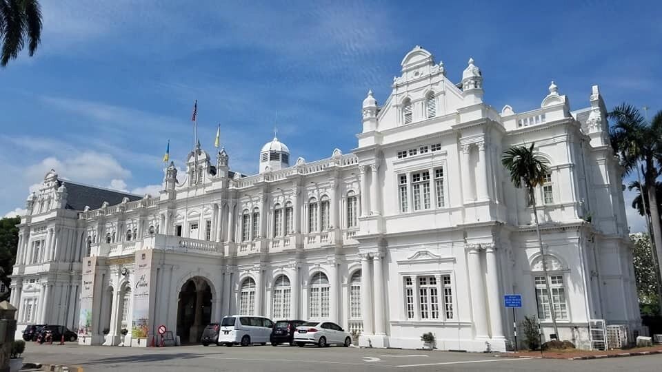 Un grand bâtiment de style colonial, de couleur blanche, doté de plusieurs dômes et de colonnes, entouré de palmiers et de voitures garées.