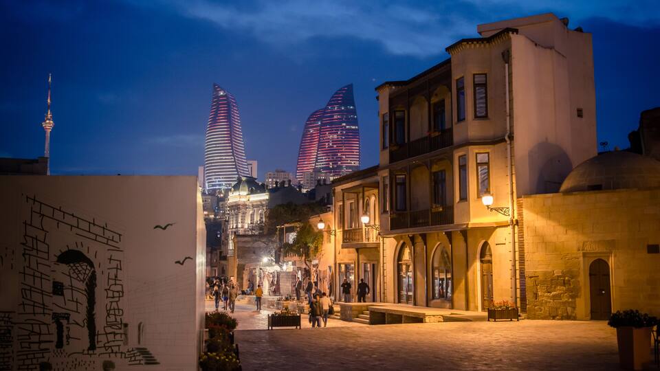 A cityscape at dusk with illuminated modern skyscrapers, historic architecture, and a bustling street.