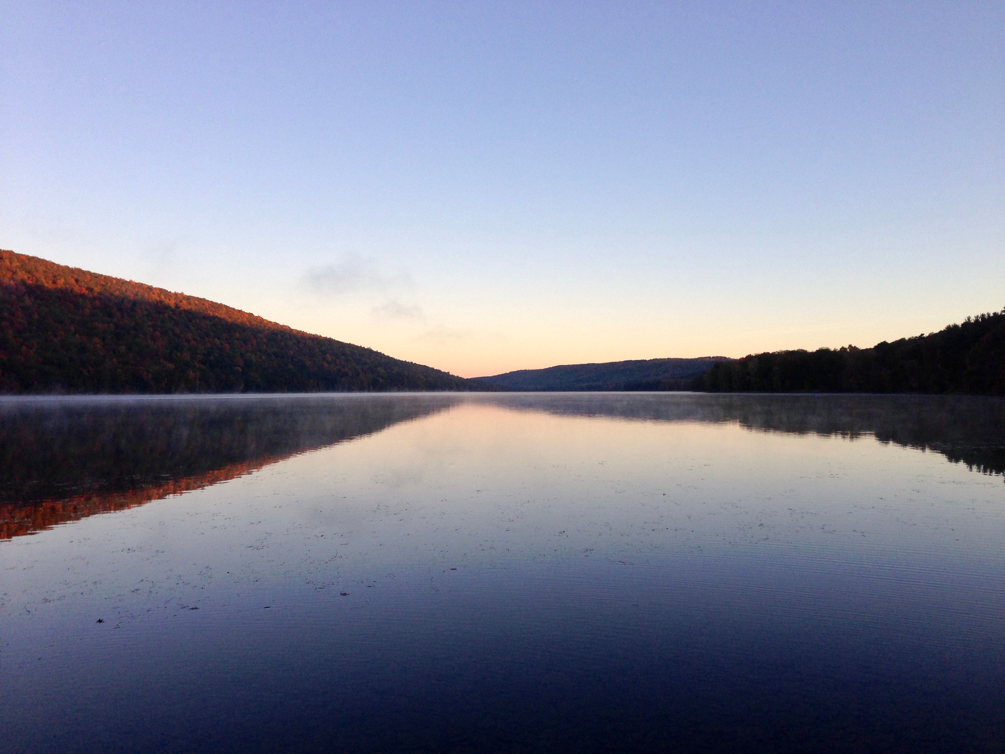A calm lake with reflections of the surrounding forest and hills.