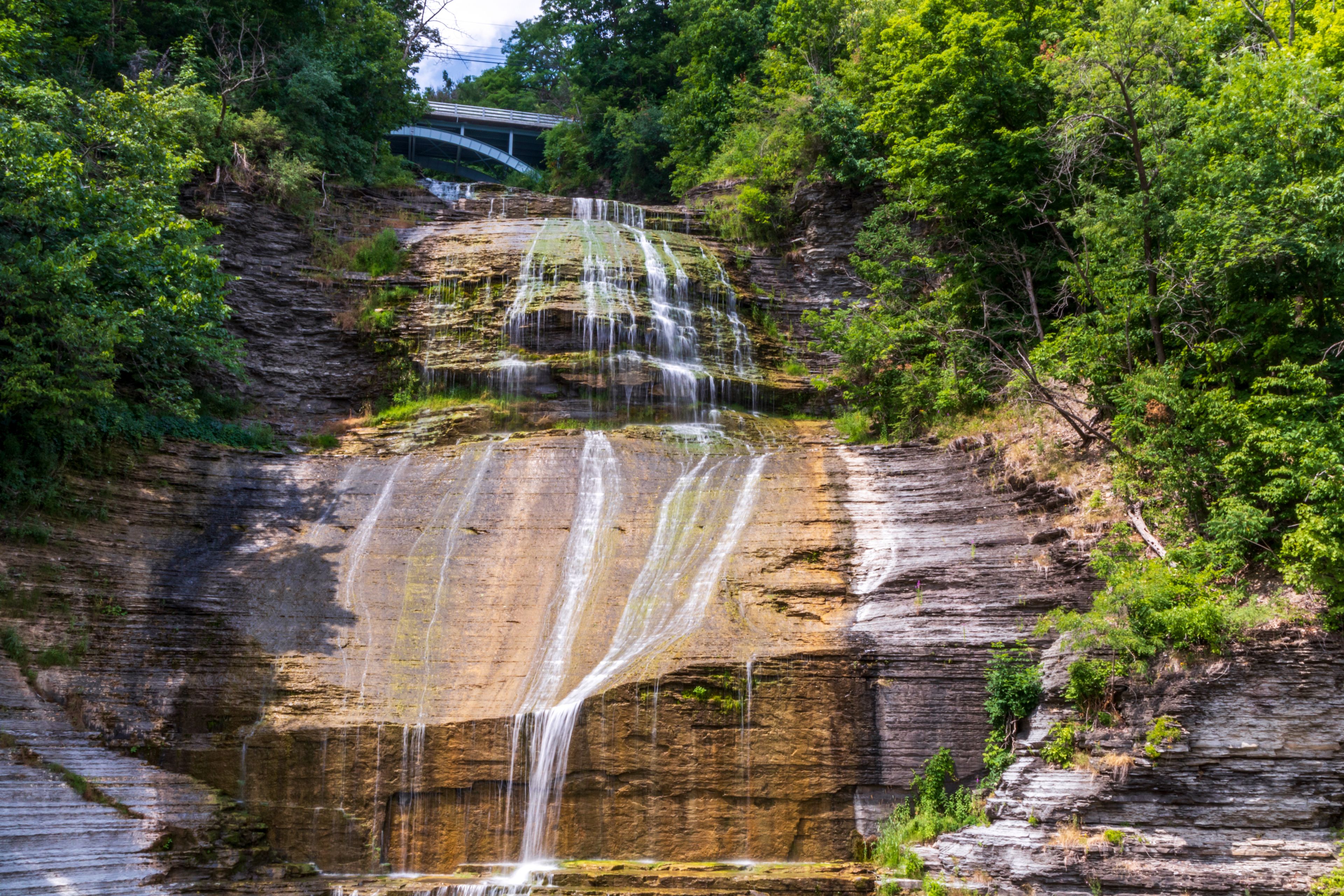 A multi-tiered waterfall cascading over layered rock formations surrounded by dense greenery.