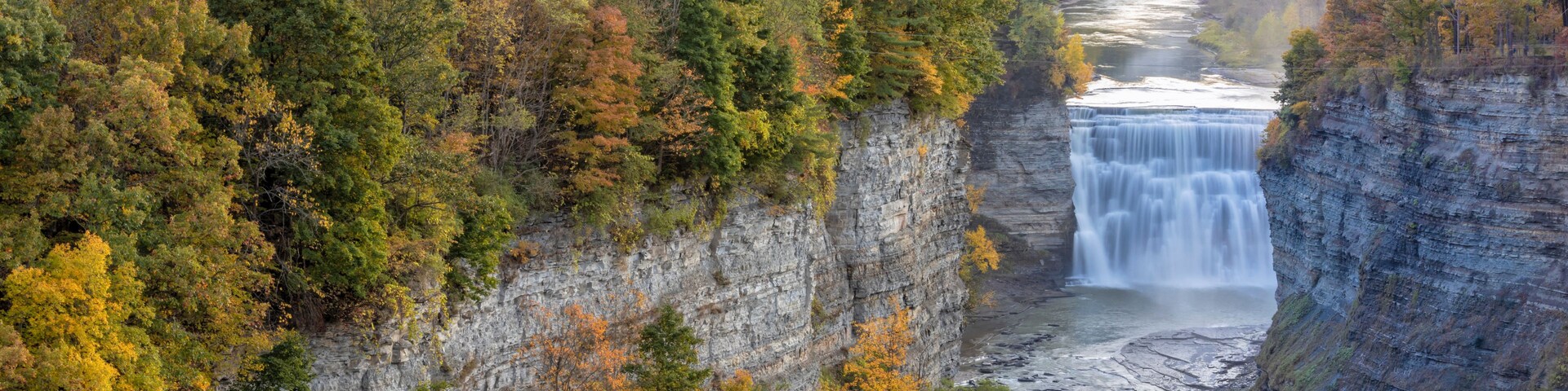 A waterfall cascading down a steep cliff, surrounded by dense forest with autumn foliage. A bridge spans across the river at the top of the cliff.