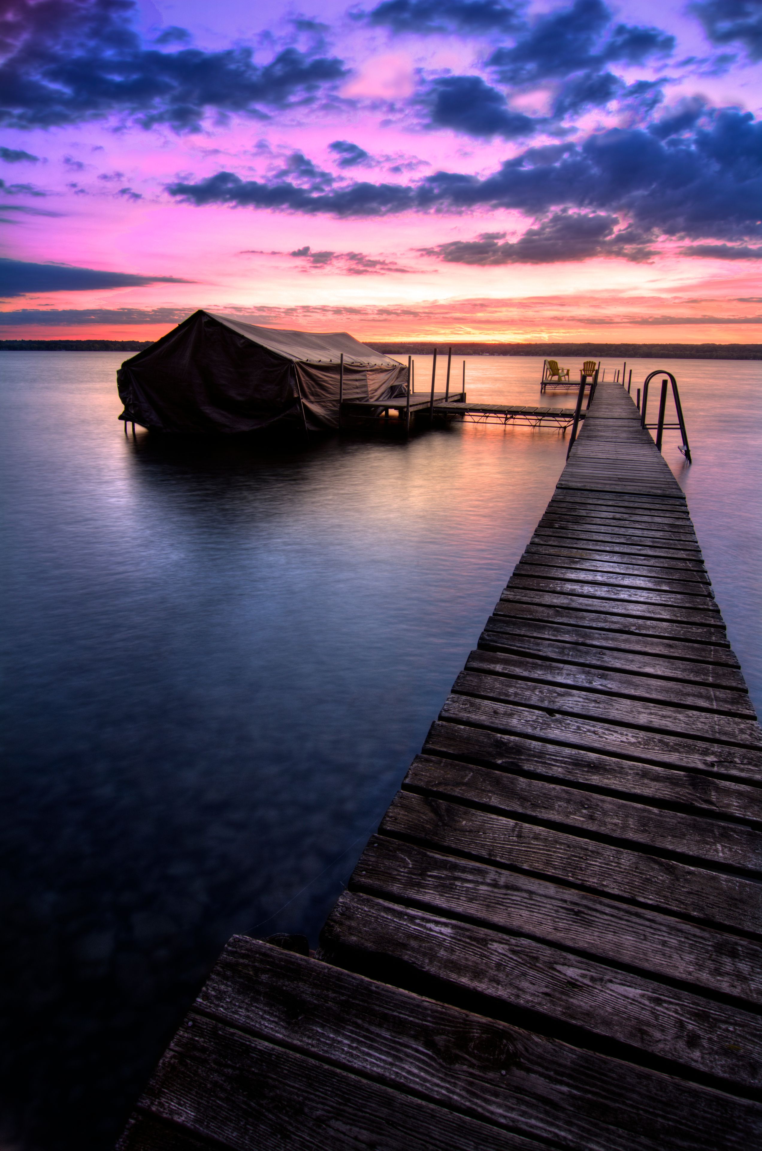 A wooden pier extending into calm water, with a covered boat at the end and a sunset sky.