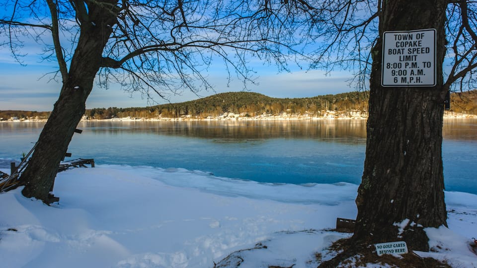 A snowy landscape with a frozen lake, a sign indicating the town of Copake boat speed limit, and a no golf carts sign.