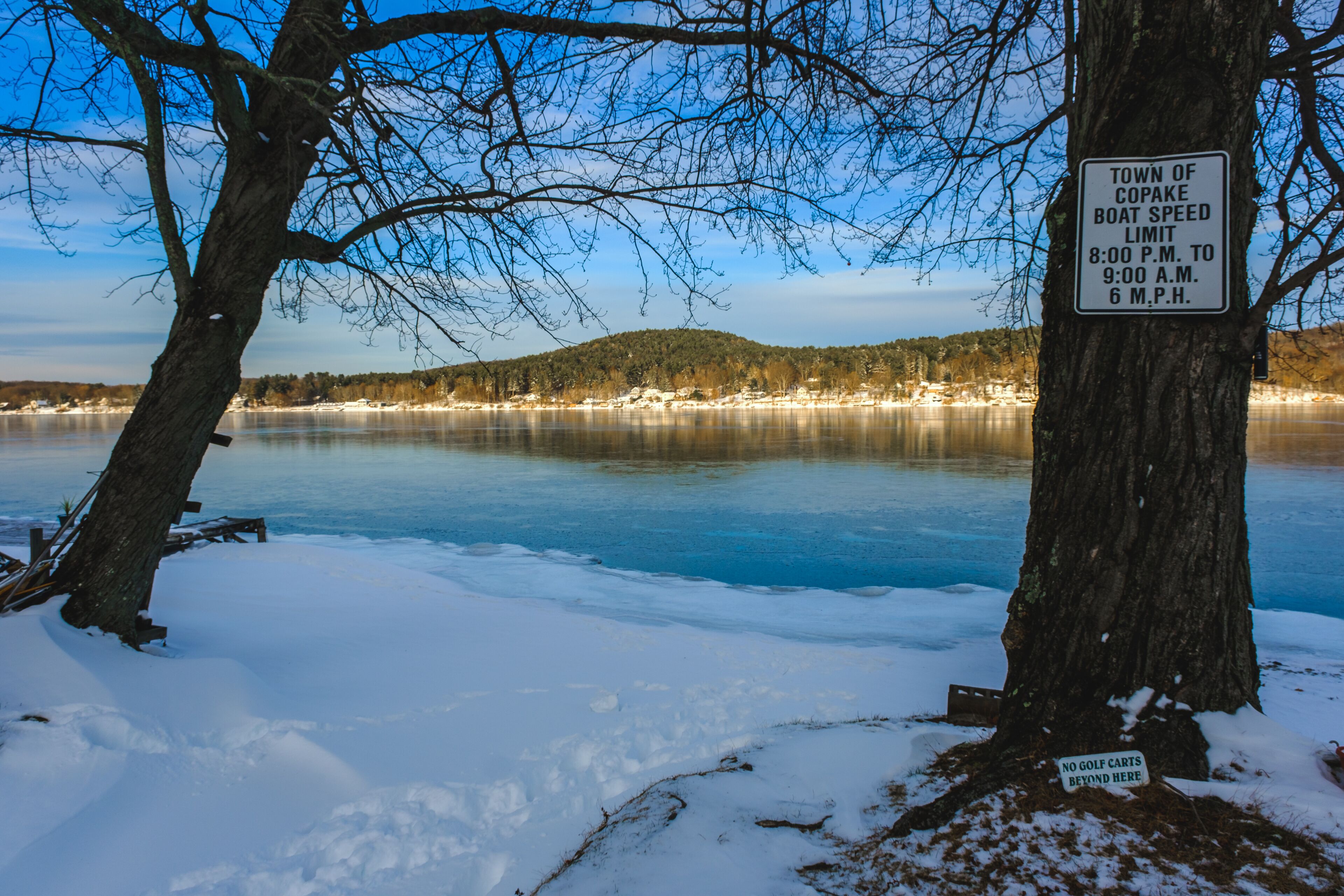 A snowy landscape with a frozen lake, a sign indicating the town of Copake boat speed limit, and a no golf carts sign.