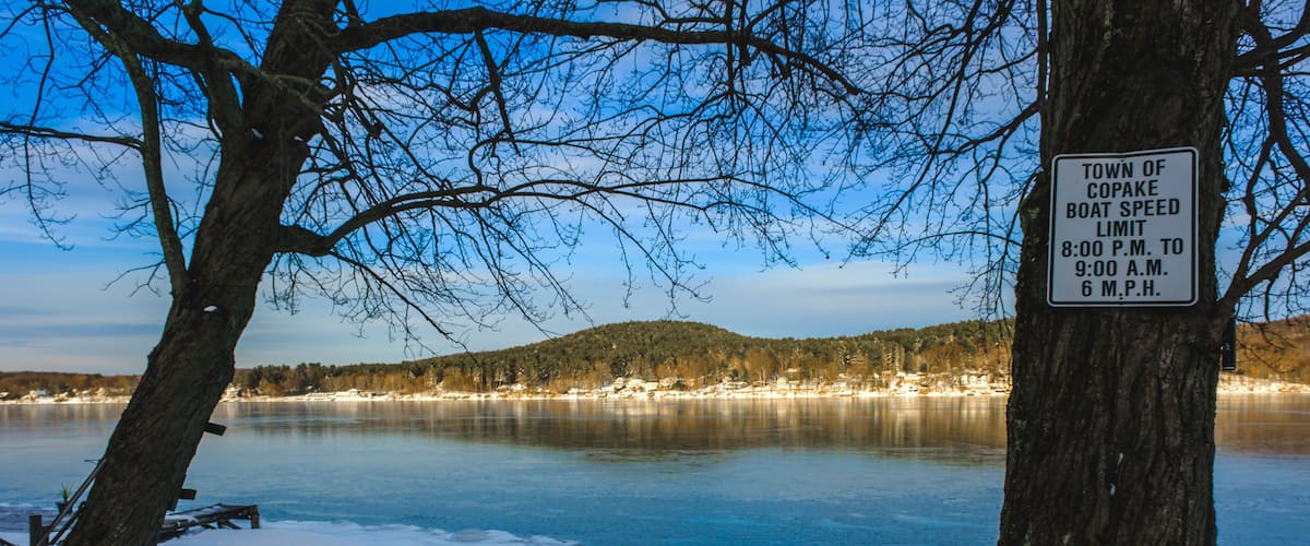 A snowy landscape with a frozen lake, a sign indicating the town of Copake boat speed limit, and a no golf carts sign.