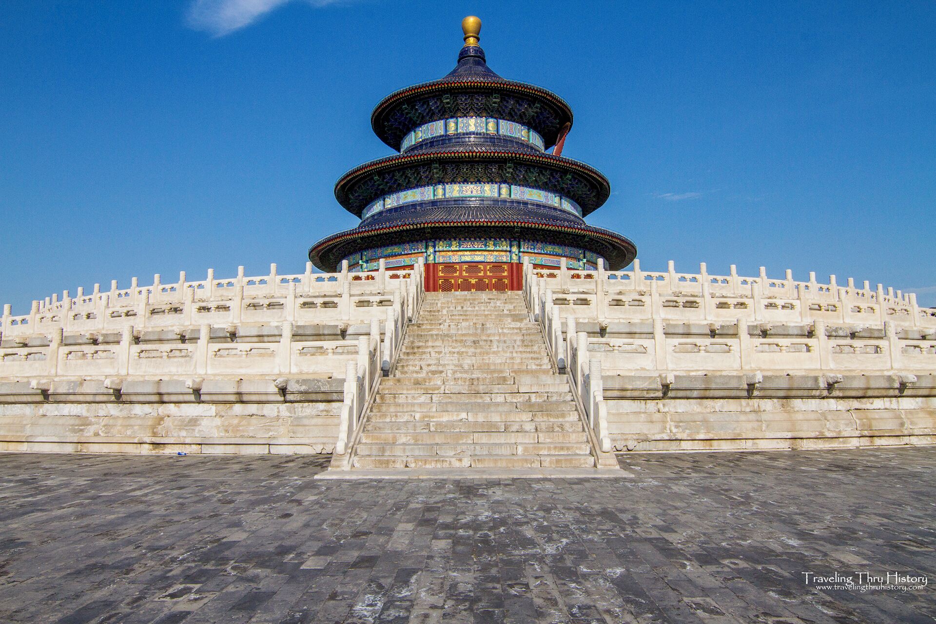 A large, multi-tiered pagoda with intricate blue and gold decorations, surrounded by a white wall with stone steps leading up to it.