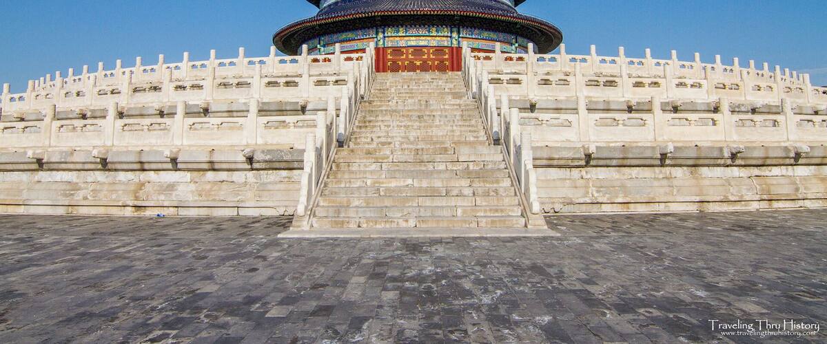 A large, multi-tiered pagoda with intricate blue and gold decorations, surrounded by a white wall with stone steps leading up to it.