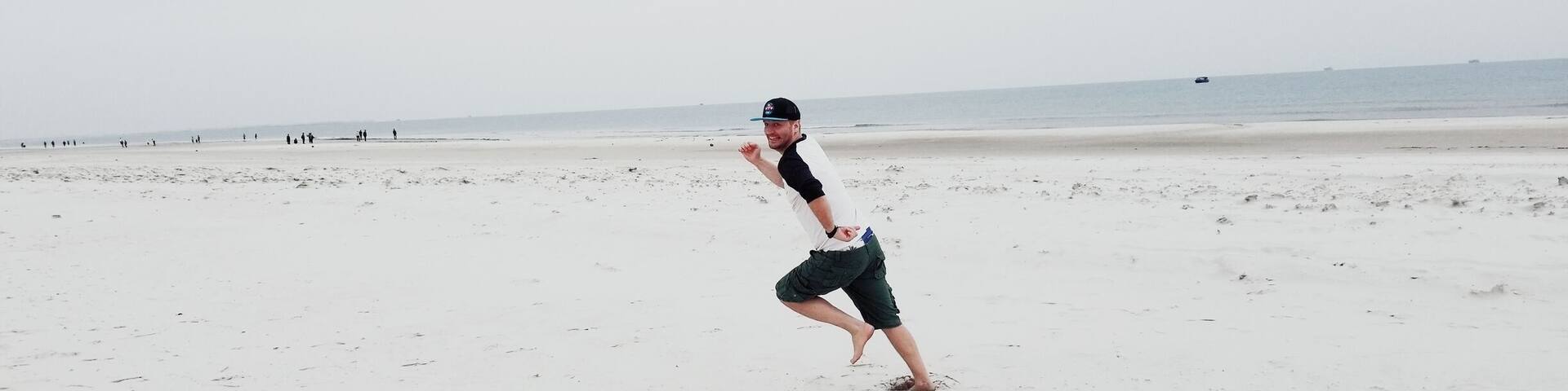 A person running on a sandy beach with a few flip-flops and a cap on the ground.