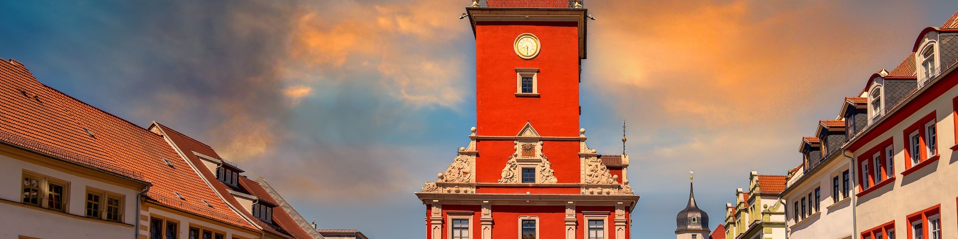 Une tour de l’horloge rouge, surmontée d’un dôme, est entourée de bâtiments historiques aux façades rouges et blanches.