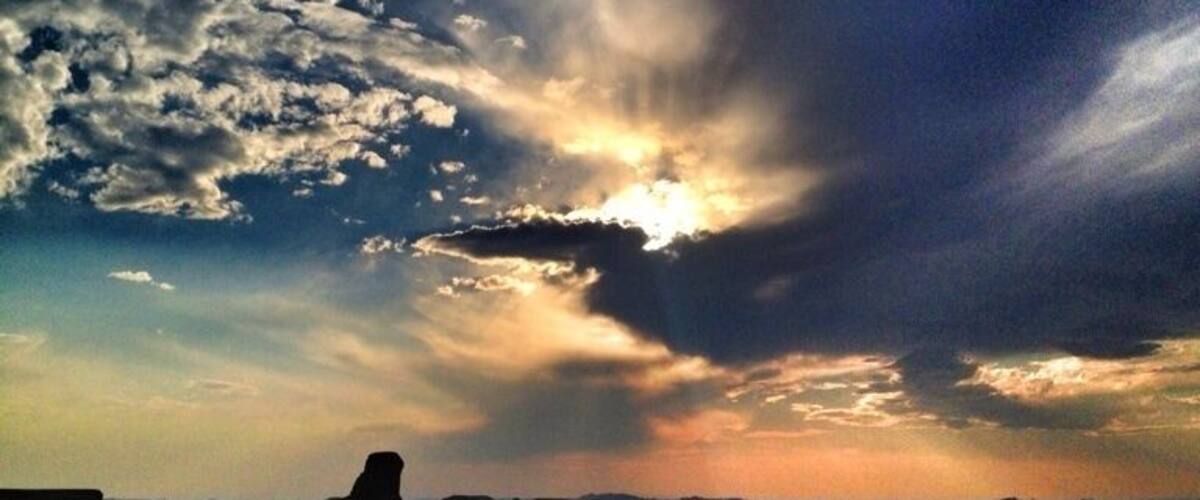 A desert landscape with a prominent rock formation silhouetted against a dramatic sky with clouds.