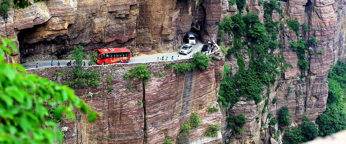 A red bus on a narrow road between cliffs, with people walking on the ledge above.
