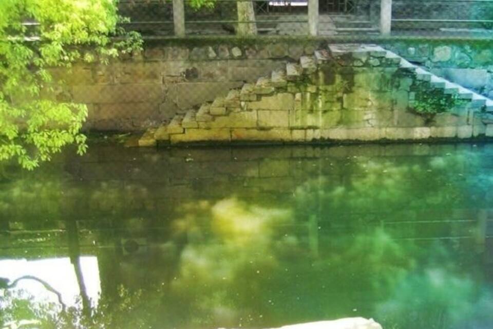 Eine Steinkonstruktion überspannt einen ruhigen Fluss, umgeben von üppiger Vegetation und einer Steinmauer.