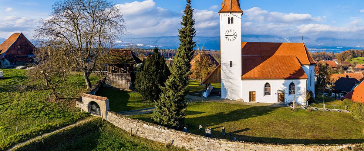 A white church with a tall steeple, surrounded by a stone wall, a wooden bridge, and a mountainous landscape in the background.