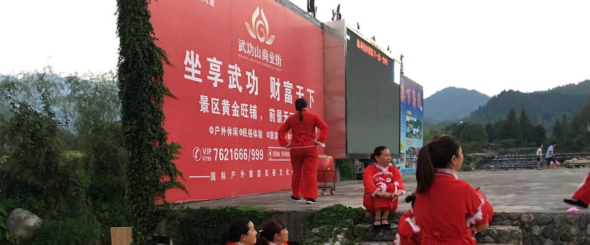 A group of people in red outfits sitting and standing near a river, with a large red sign in the background.
