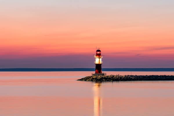 Ein Leuchtturm auf einem Felsen bei Sonnenuntergang, ruhige Gewässer spiegeln den Himmel wider.