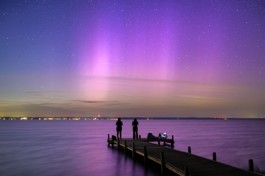 A night scene with a wooden pier extending into a calm body of water, under a starry sky with visible auroras.