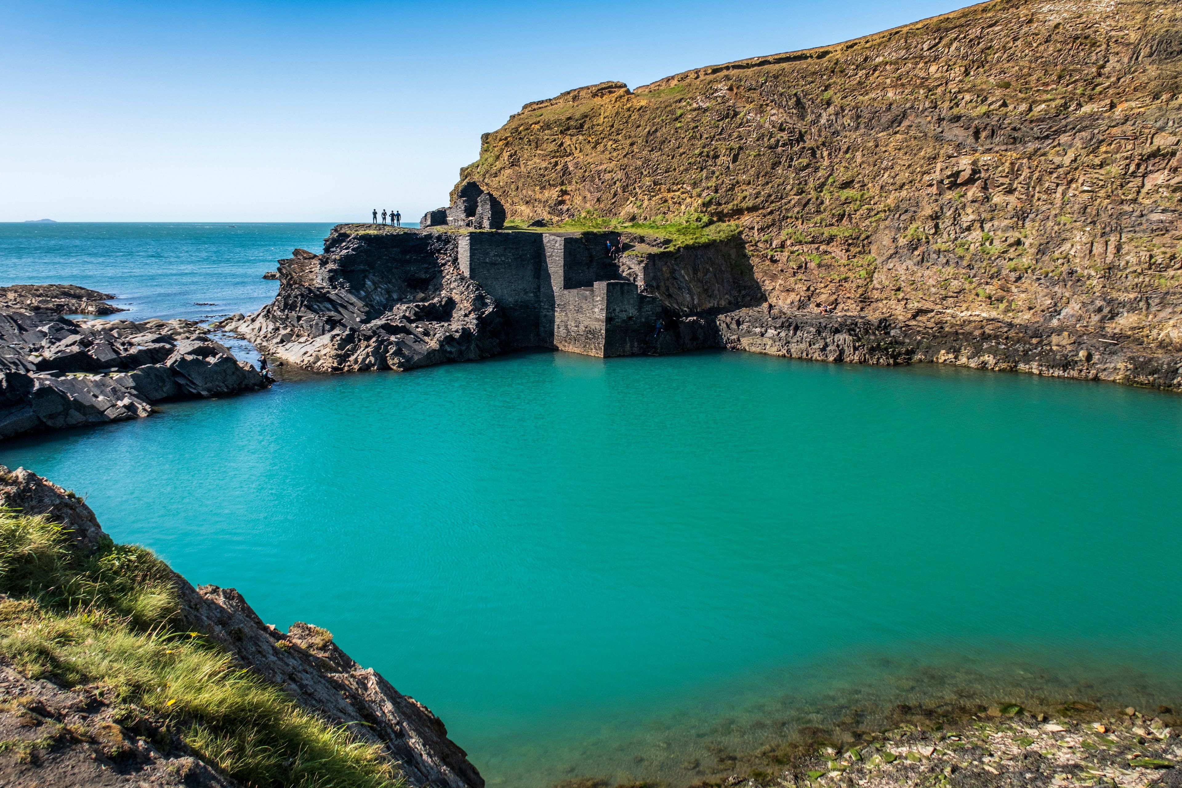 A coastal landscape with a rocky cliff, a clear turquoise sea, and a small group of people on a cliff edge.