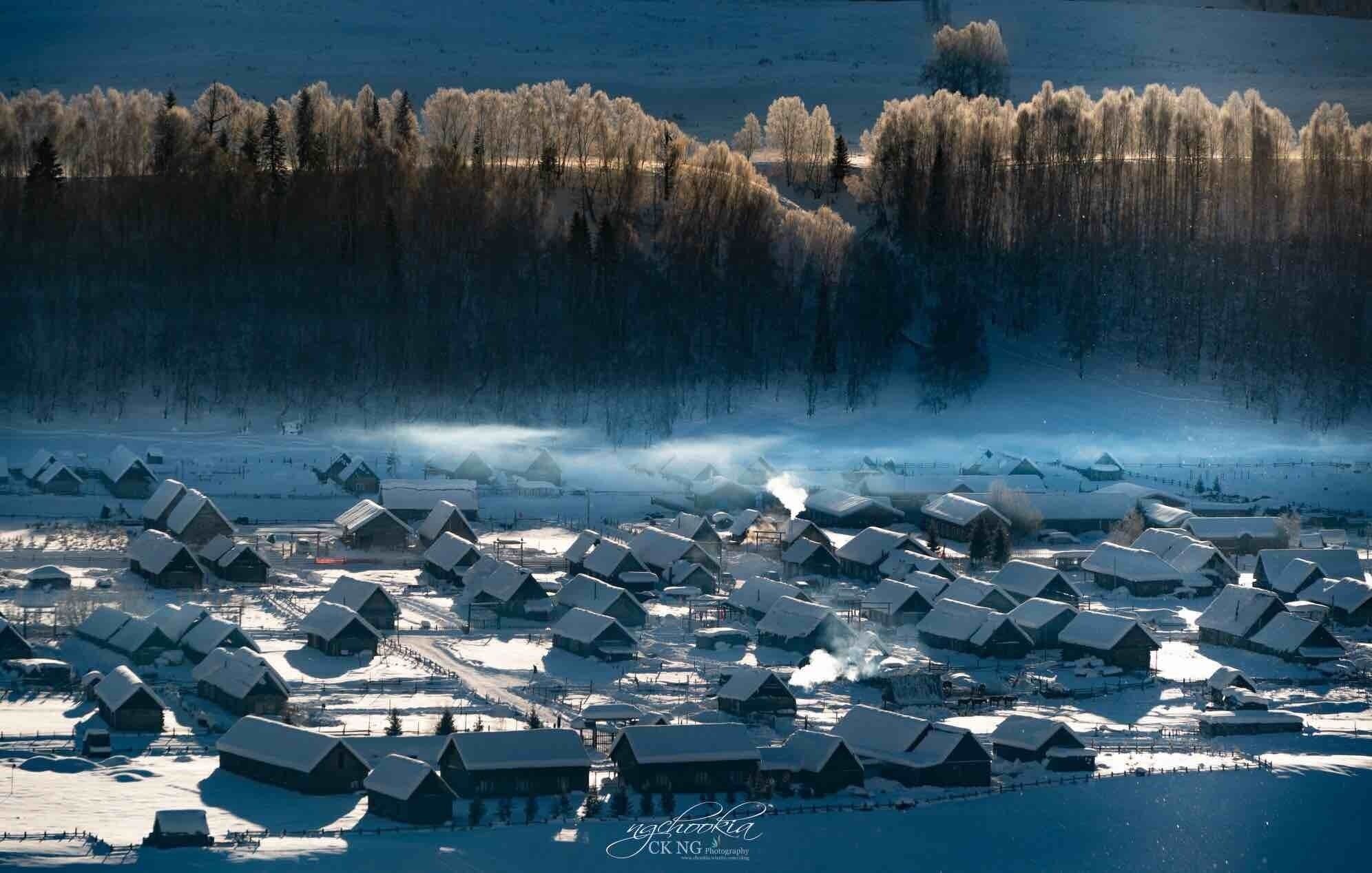 A snow-covered village with wooden houses and a forest in the background.