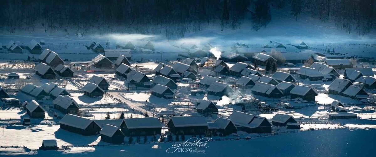 A snow-covered village with wooden houses and a forest in the background.
