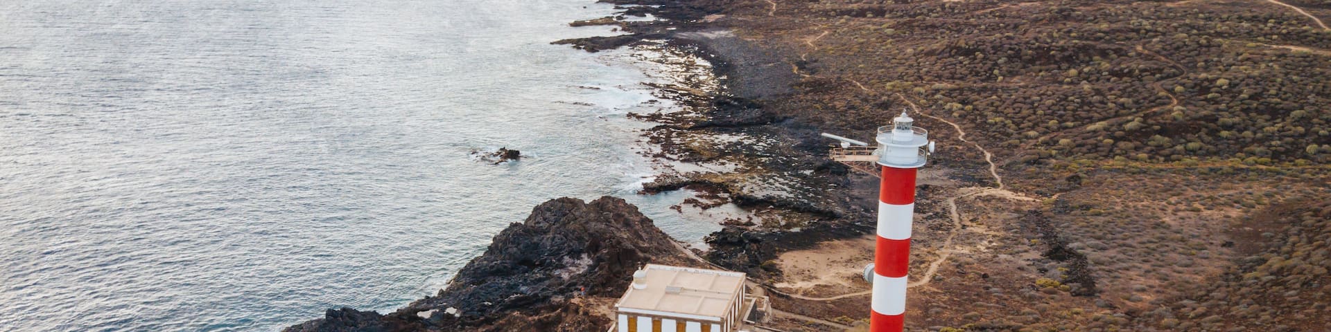 A coastal landscape with a prominent red and white lighthouse, rocky terrain, and a clear view of the ocean.