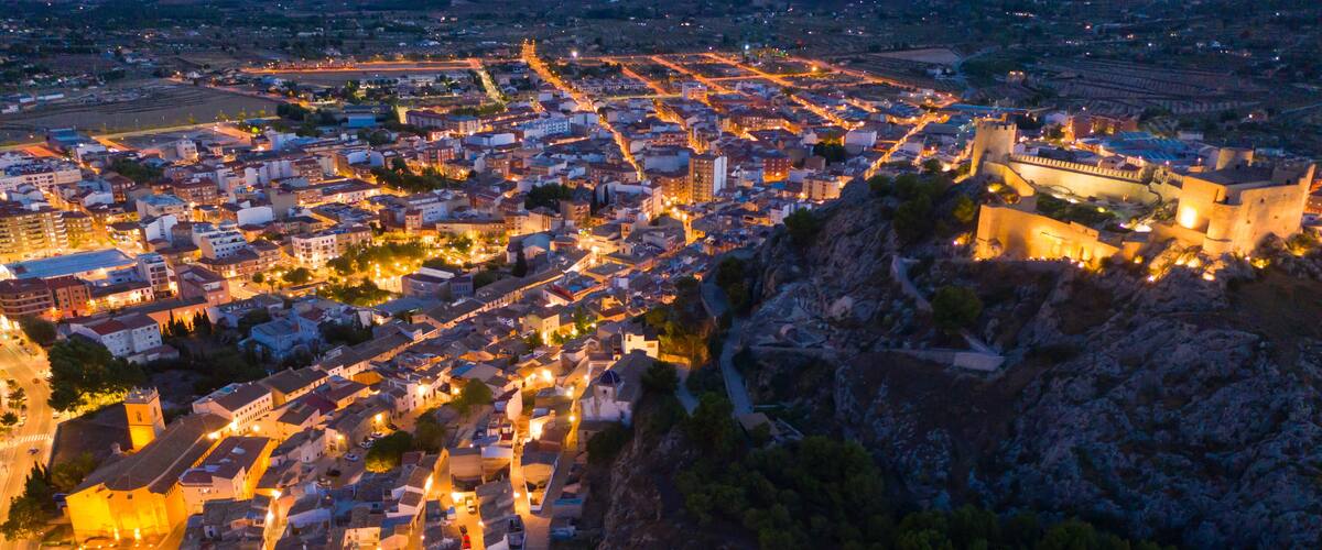 Aerial view of medieval fortified Castalla Castle on top of stone hill on background with townscape in twilight, province of Alicante, Spain..