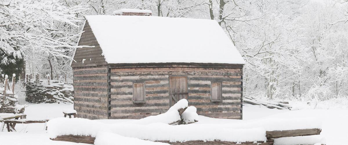 A snow-covered log cabin surrounded by snow-covered trees and logs.