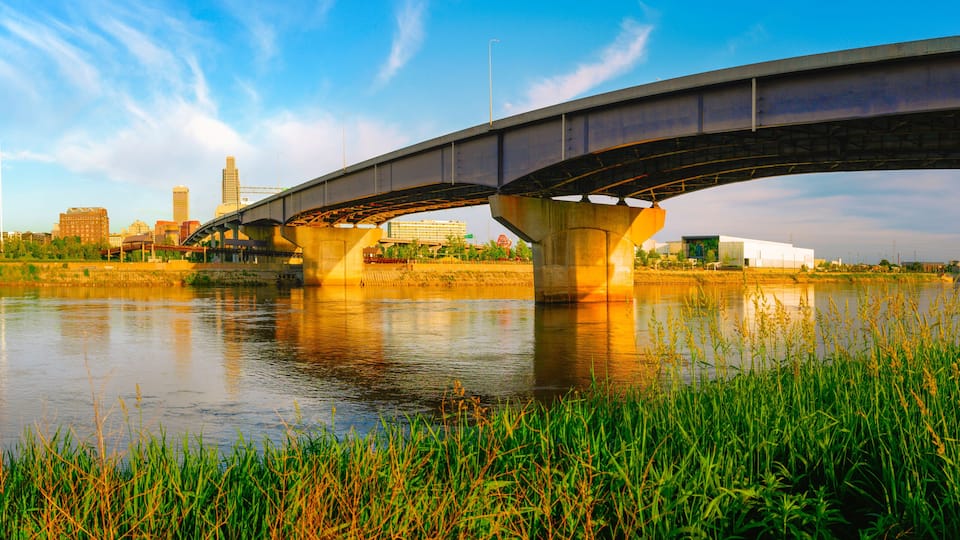 Omaha City Skyline, I-480 Missouri River Bridge, and gentle water flow at warm glowing sunrise: A tranquil summer landscape from Council Bluffs Riverfront in Iowa