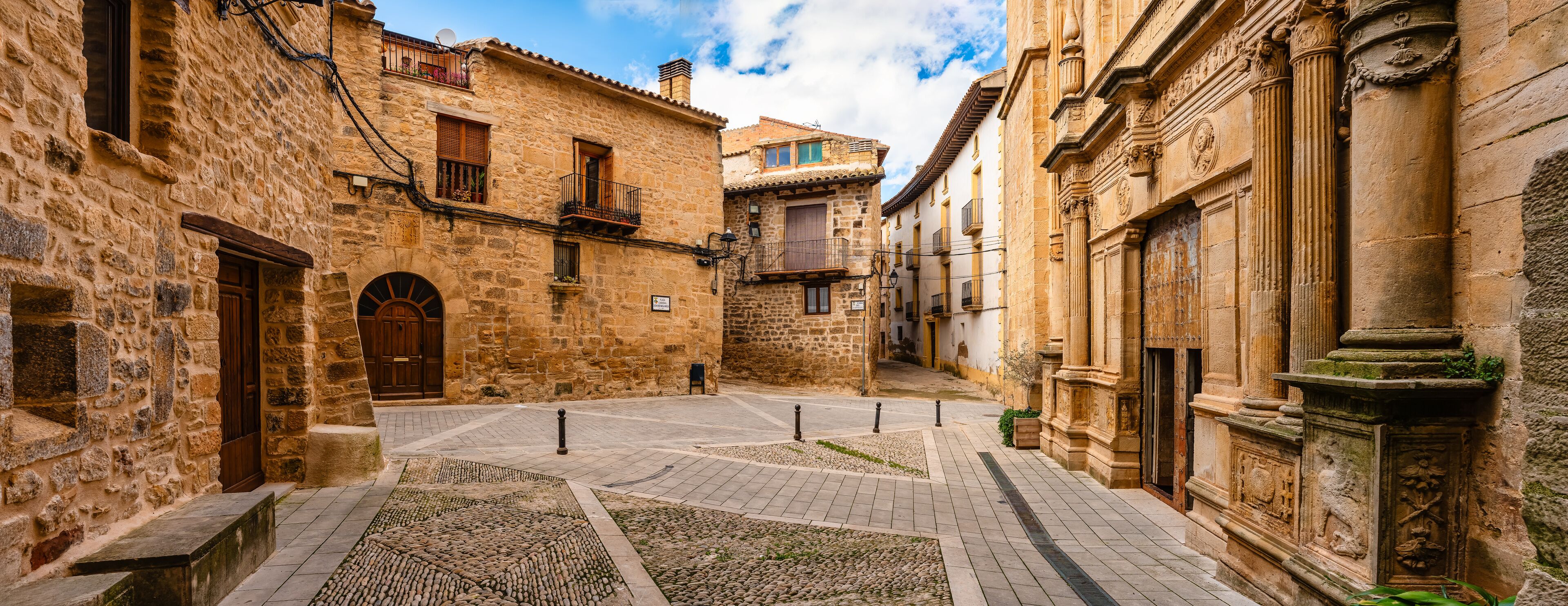 A narrow cobblestone street flanked by historic stone buildings with arched doorways and balconies.