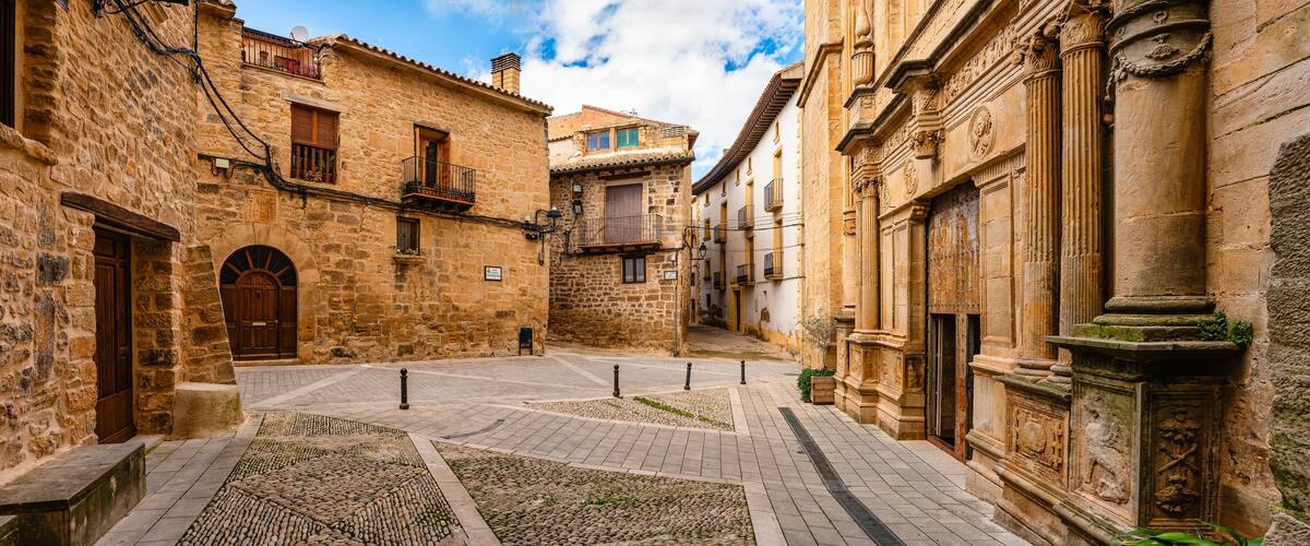 A narrow cobblestone street flanked by historic stone buildings with arched doorways and balconies.