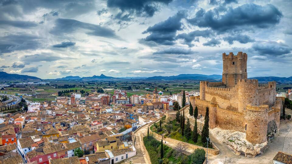A historic castle overlooking a densely built town with a mountainous backdrop.