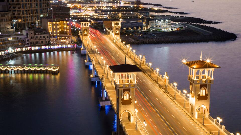 A brightly lit bridge with light trails from vehicles, a calm body of water, and a distant cityscape at dusk.