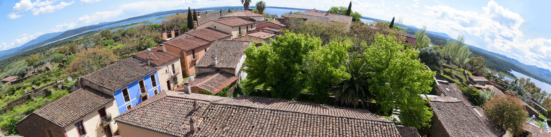 Vista panorámica de un pueblo costero con edificios de tejados de terracota, abundante vegetación y un cielo azul claro.
