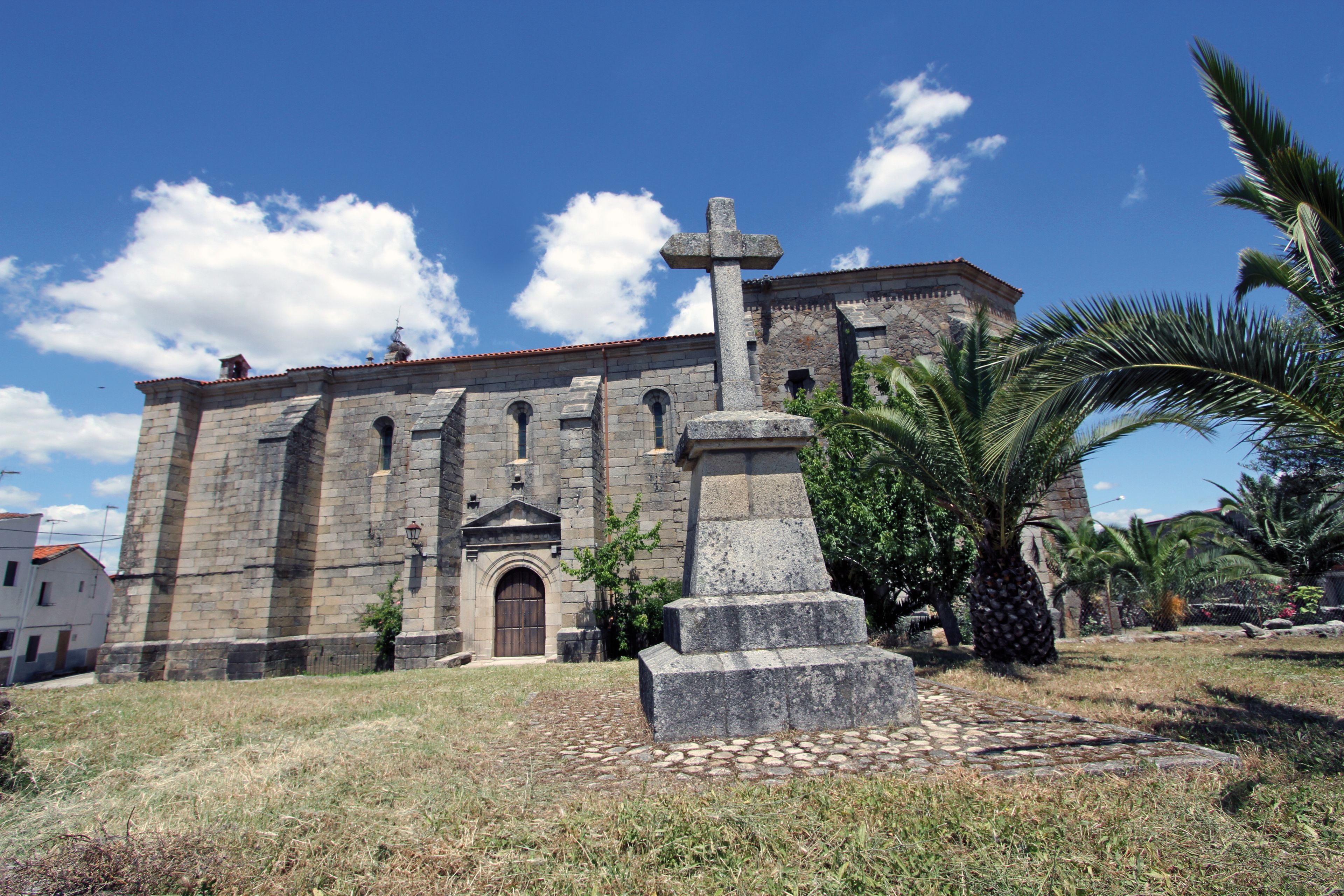 A historic stone church with a prominent cross, surrounded by palm trees and a clear blue sky.