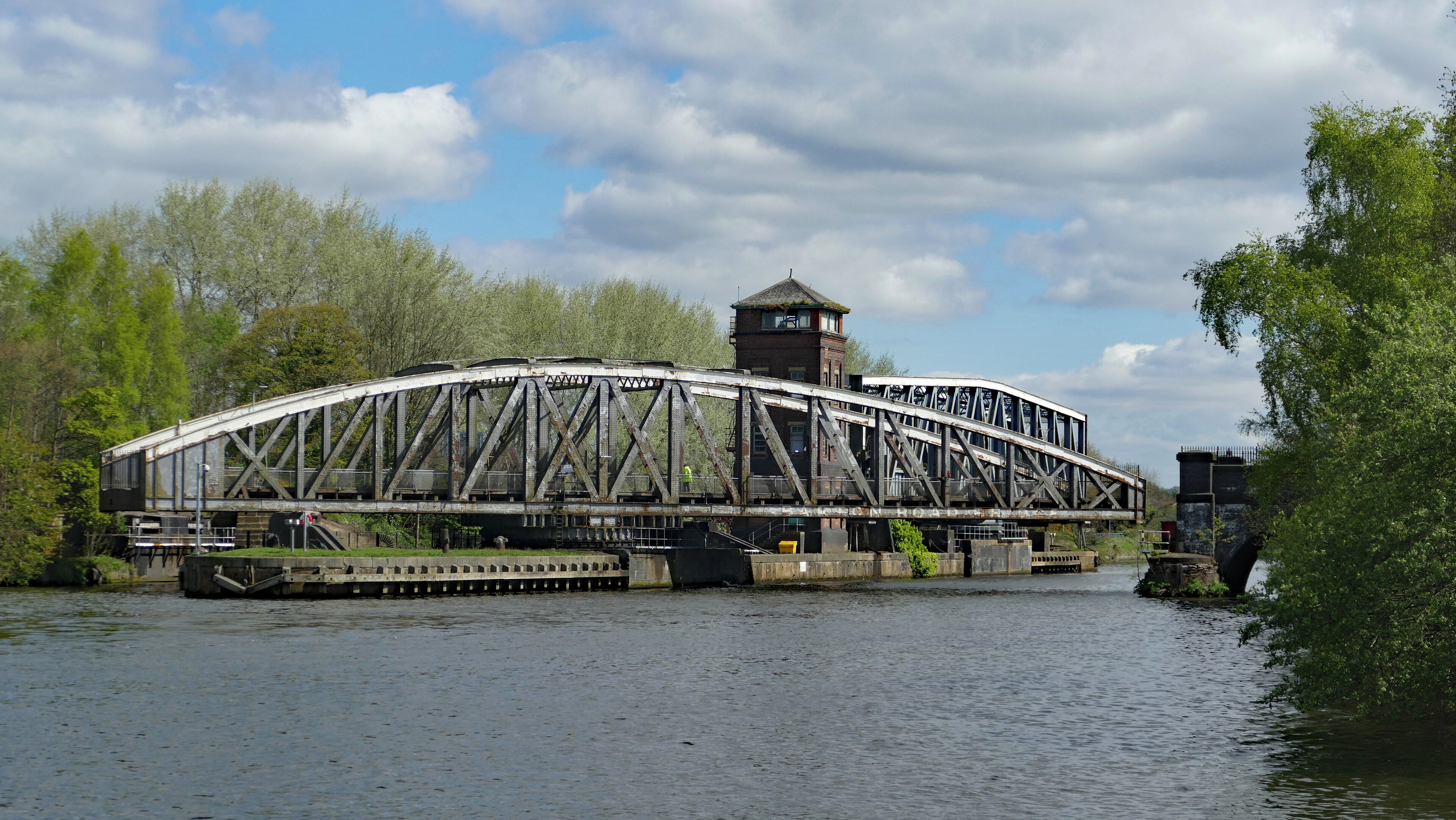 Barton Bridge, a swing bridge carrying Barton Road over the Manchester Ship Canal, turning open to allow a waiting ship to pass. In the background is the Barton Aqueduct, which is also a swing bridge, and at the time of the photo had already turned fully open in readiness for the ship. This view is from the southwest. Additional information from the photographer's original post to Flickr: The aqueduct was shifted about fifteen minutes before the road bridge meaning it's easy to catch both operations. This is from the only decent vantage point on the canal side just off the Old Barton Road (which provides decent parking for anyone driving to view the mechanical marvels). The road bridge is opened "just in time" for any passing ships.
