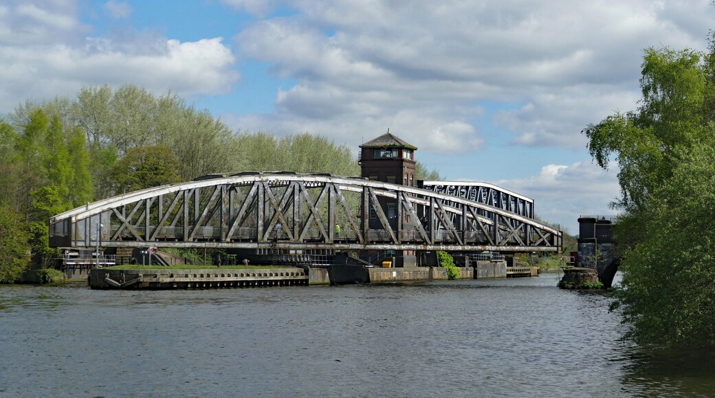 Barton Bridge, a swing bridge carrying Barton Road over the Manchester Ship Canal, turning open to allow a waiting ship to pass. In the background is the Barton Aqueduct, which is also a swing bridge, and at the time of the photo had already turned fully open in readiness for the ship. This view is from the southwest. Additional information from the photographer's original post to Flickr: The aqueduct was shifted about fifteen minutes before the road bridge meaning it's easy to catch both operations. This is from the only decent vantage point on the canal side just off the Old Barton Road (which provides decent parking for anyone driving to view the mechanical marvels). The road bridge is opened "just in time" for any passing ships.