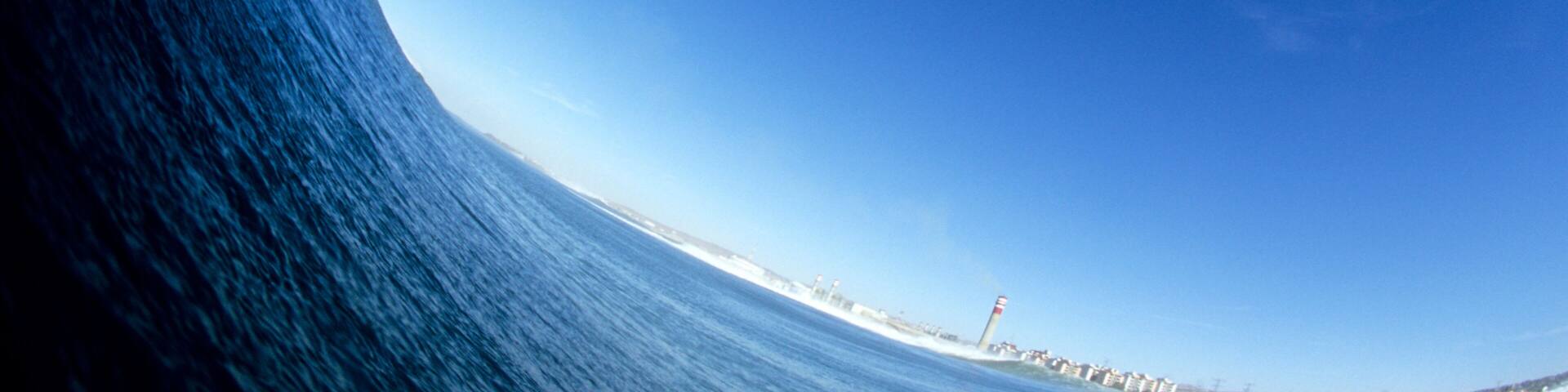 A large wave curling over the ocean with a clear blue sky and a distant lighthouse.