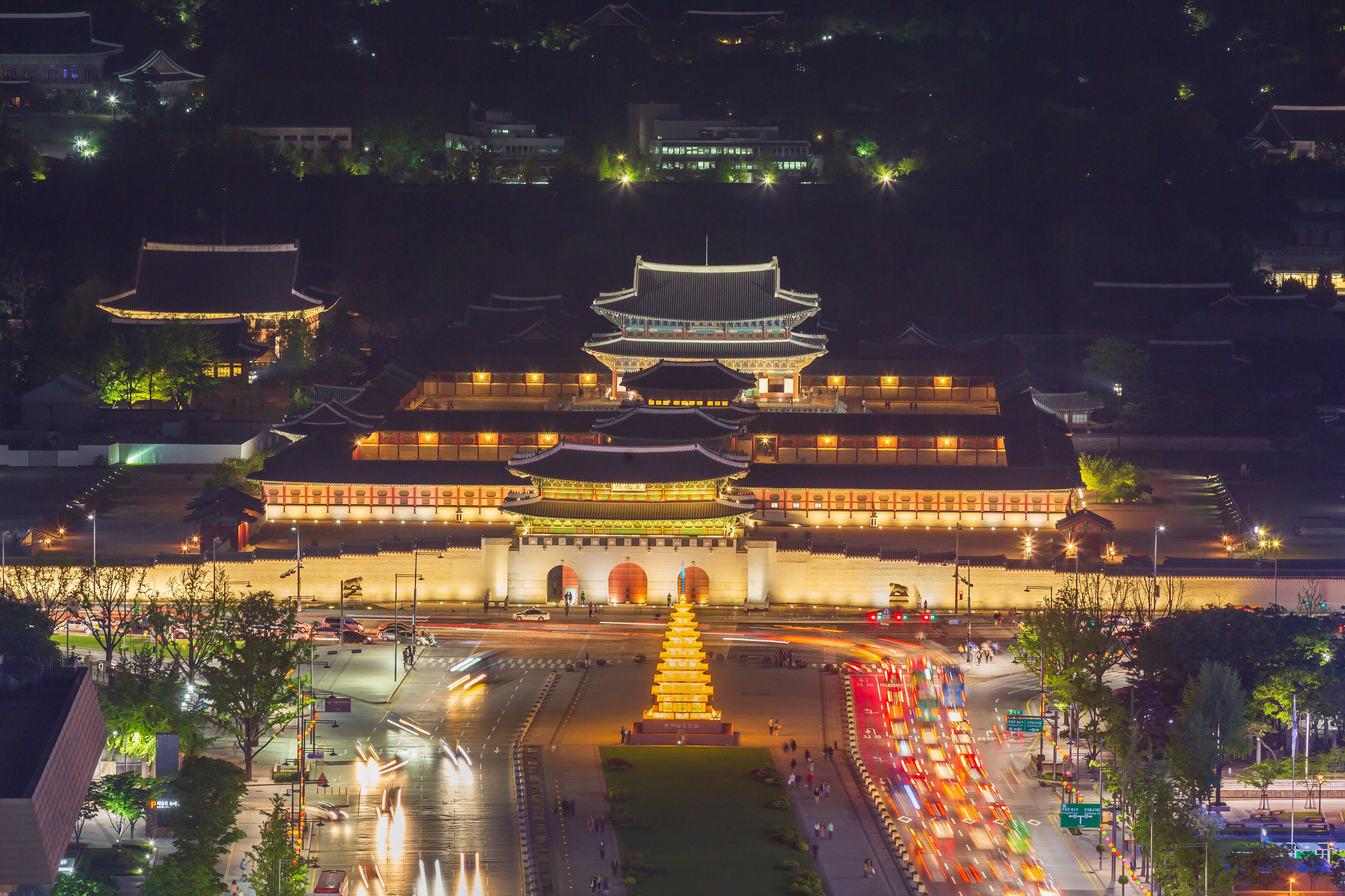 A traditional multi-tiered building with illuminated rooftops, surrounded by a moat and a busy street with light trails from moving vehicles.
