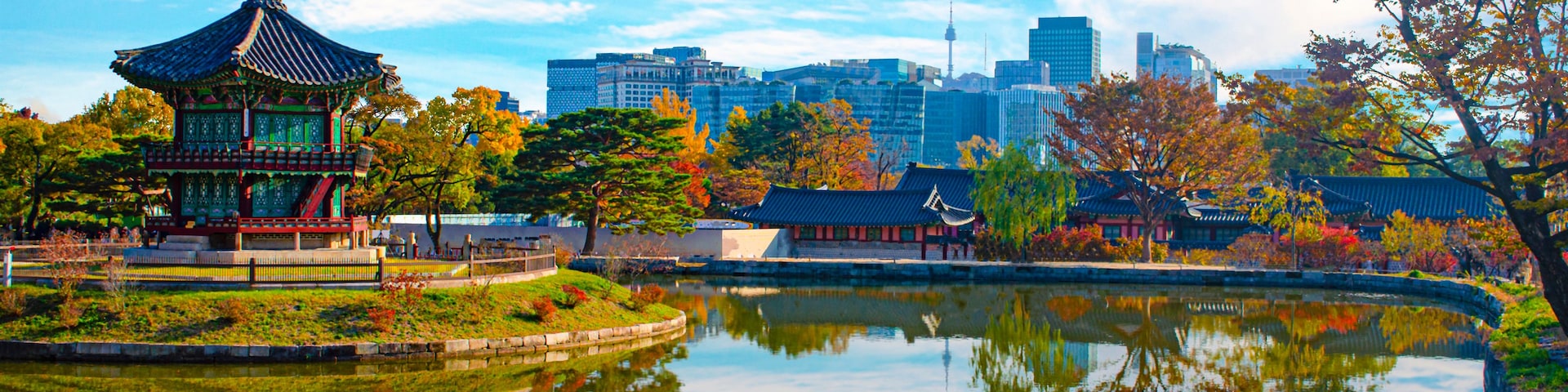 Um pavilhão coreano tradicional à beira de um lago tranquilo, refletindo a estrutura e as árvores ao redor. Ao fundo, edifícios modernos e um céu azul límpido.