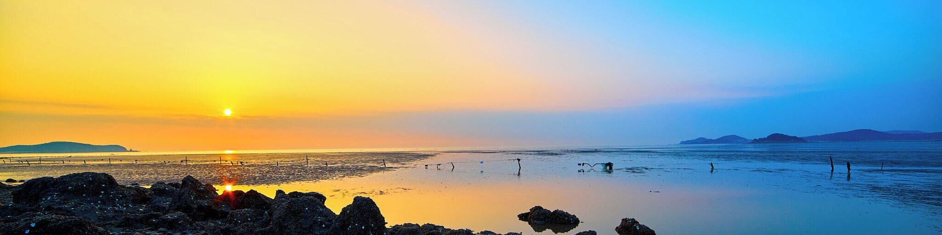 Um pôr do sol sobre uma costa rochosa, com águas calmas refletindo o céu.