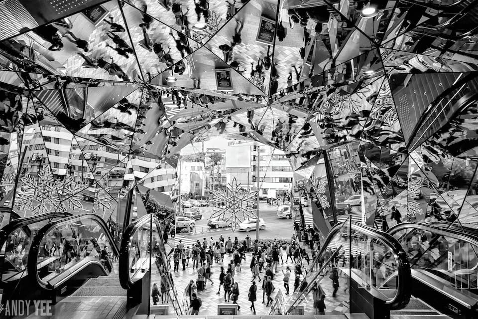 Un centre comercial animat, amb escales mecàniques, sostres de vidre i reflexos de persones i objectes.